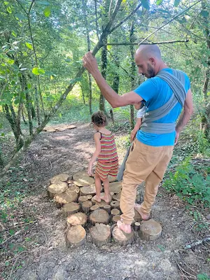 Adulte portant un bébé en écharpe et enfant marchant sur des rondins de bois sur le sentier pieds nus du Domaine de Larrous – Domaine de Larrous, Gaujacq (Landes)
